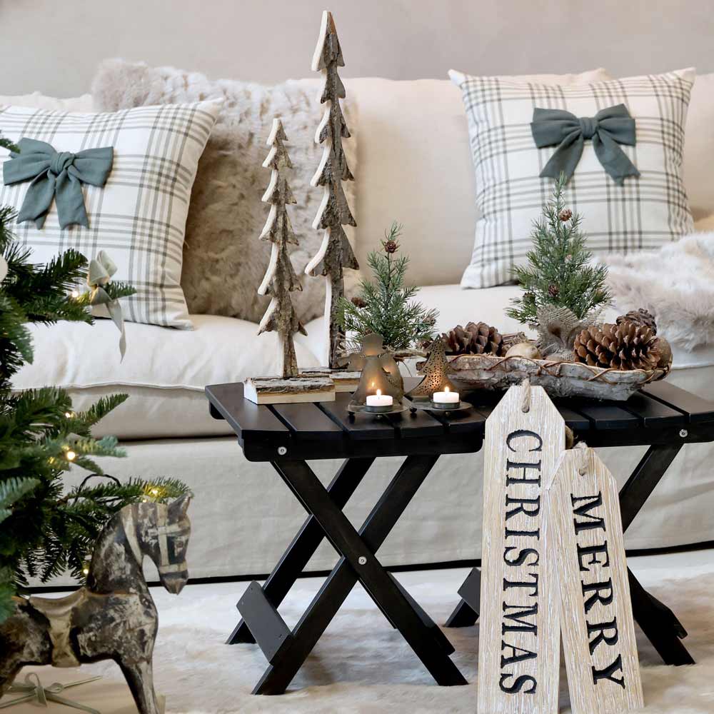 Eine gemütliche Wohnzimmerszene besteht aus einem Sofa mit karierten Kissen, Weihnachtsbäumen auf dem Tisch, darunter dem Chic Antique - Fleur Tannenbaum mit Zapfen 25 cm, brennenden Kerzen, Tannenzapfen und einem „Frohe Weihnachten“-Schild auf einem schwarzen Klapptisch. Im Vordergrund steht ein Spielzeugpferd.