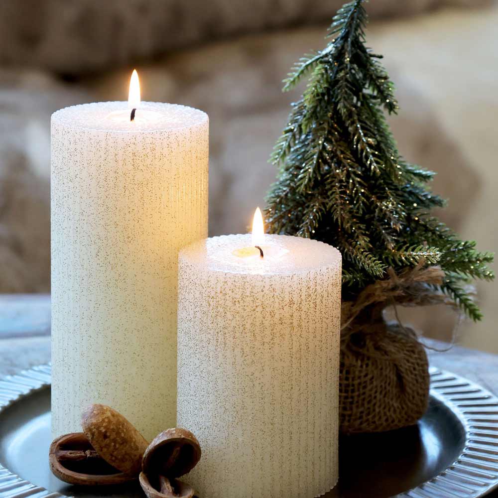 Neben dem Chic Antique – Fleur Tannenbaum mit Glitzergold stehen zwei brennende weiße Kerzen auf einer metallischen Oberfläche.