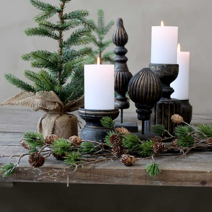 Ein kleiner Weihnachtsbaum im Topf und brennende weiße Kerzen auf einem rustikalen Holztisch, geschmückt mit dem Chic Antique Fleur Zedernzweig mit Zapfen 96 cm in Grün.