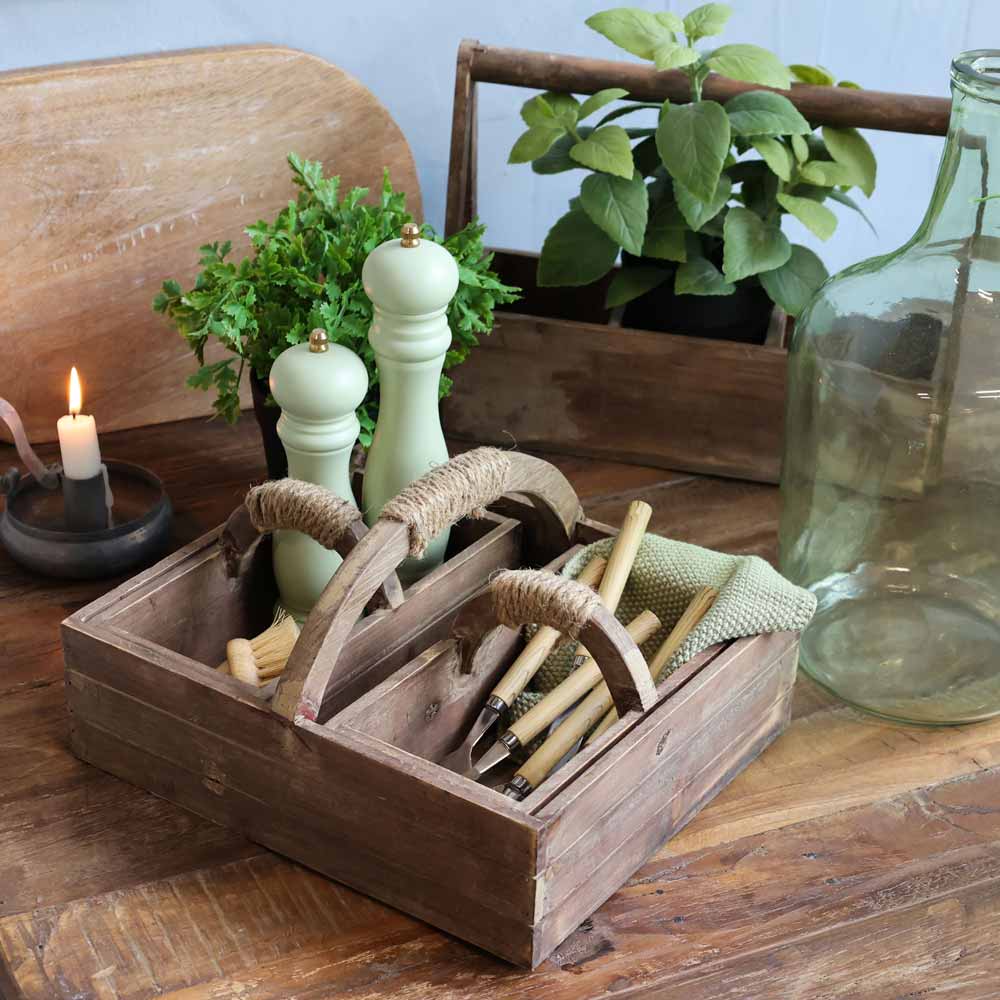 Der Chic Antique - Kasten mit Handgriff und 2 Kisten enthält Holzlöffel, ein grünes Tuch sowie Salz- und Pfeffermühlen. In der Nähe stehen eine brennende Kerze, Topfpflanzen und eine große Glasflasche auf dem Holztisch.