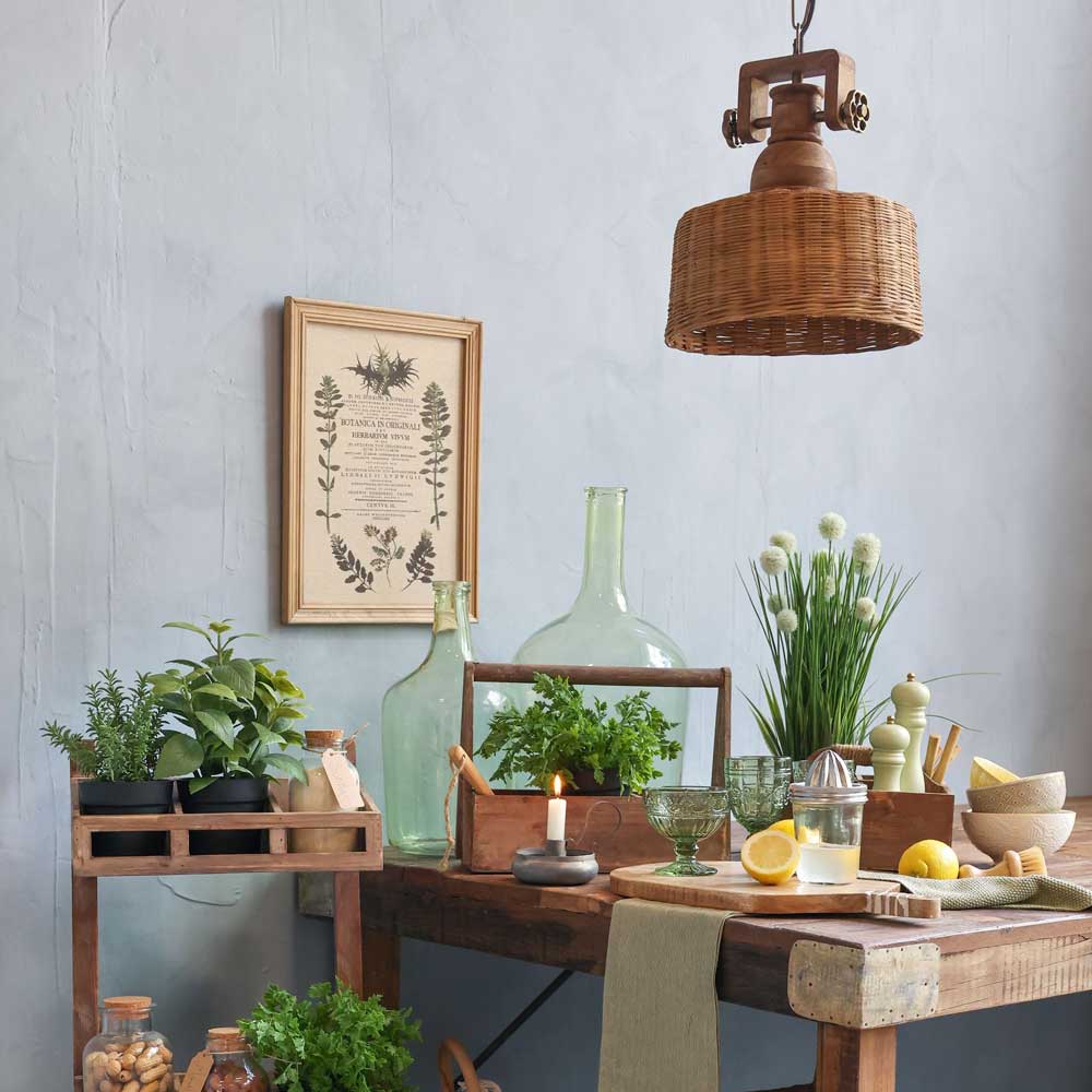 Rustikale Kücheneinrichtung mit Chic Antiques Caddy mit Griff und drei Fächern, einem Holztisch, Topfpflanzen, Glasflaschen, einer Kerze, Zitronen und Kräutern. Darüber hängt eine Korblampe; ein gerahmter botanischer Druck verstärkt den Charme.