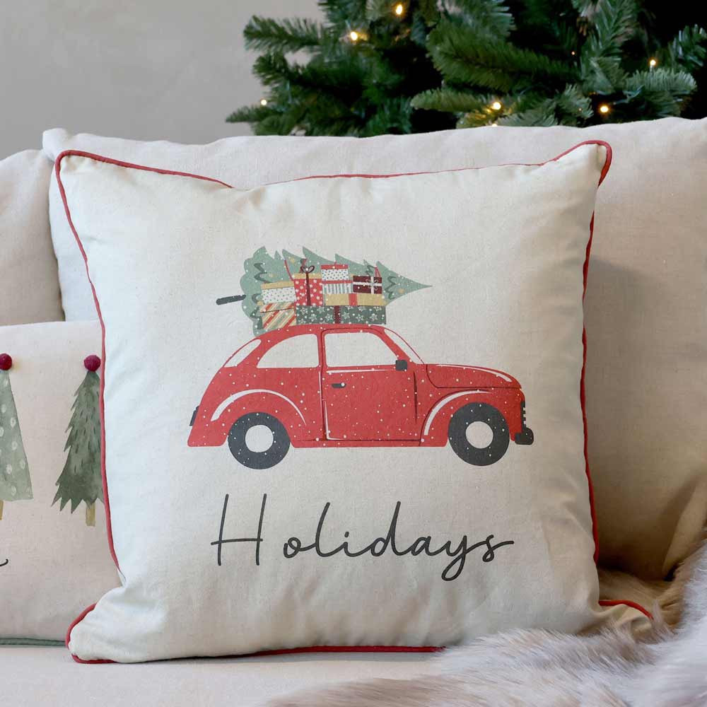 Der Chic Antique - Kissenbezug Weihnachtsauto Leinen 50 x 50 cm zeigt ein rotes Auto mit einem Weihnachtsbaum auf dem Dach und darunter das Wort „Holidays“. Im Hintergrund sind ein weiteres Kissen und ein Weihnachtsbaum zu sehen.