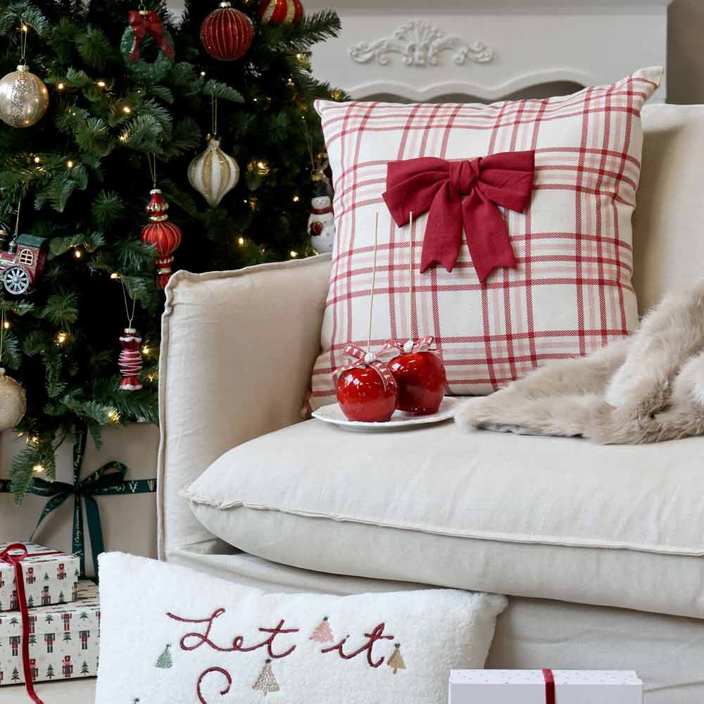 Ein dekoriertes Wohnzimmer mit einem Weihnachtsbaum, einem karierten Kissen von Chic Antique in Kirsche mit einer Schleife auf dem Sofa, dazu ein weißes Kissen mit der Aufschrift „Let it Snow“ und auf einem Teller arrangierte rote Ornamente.