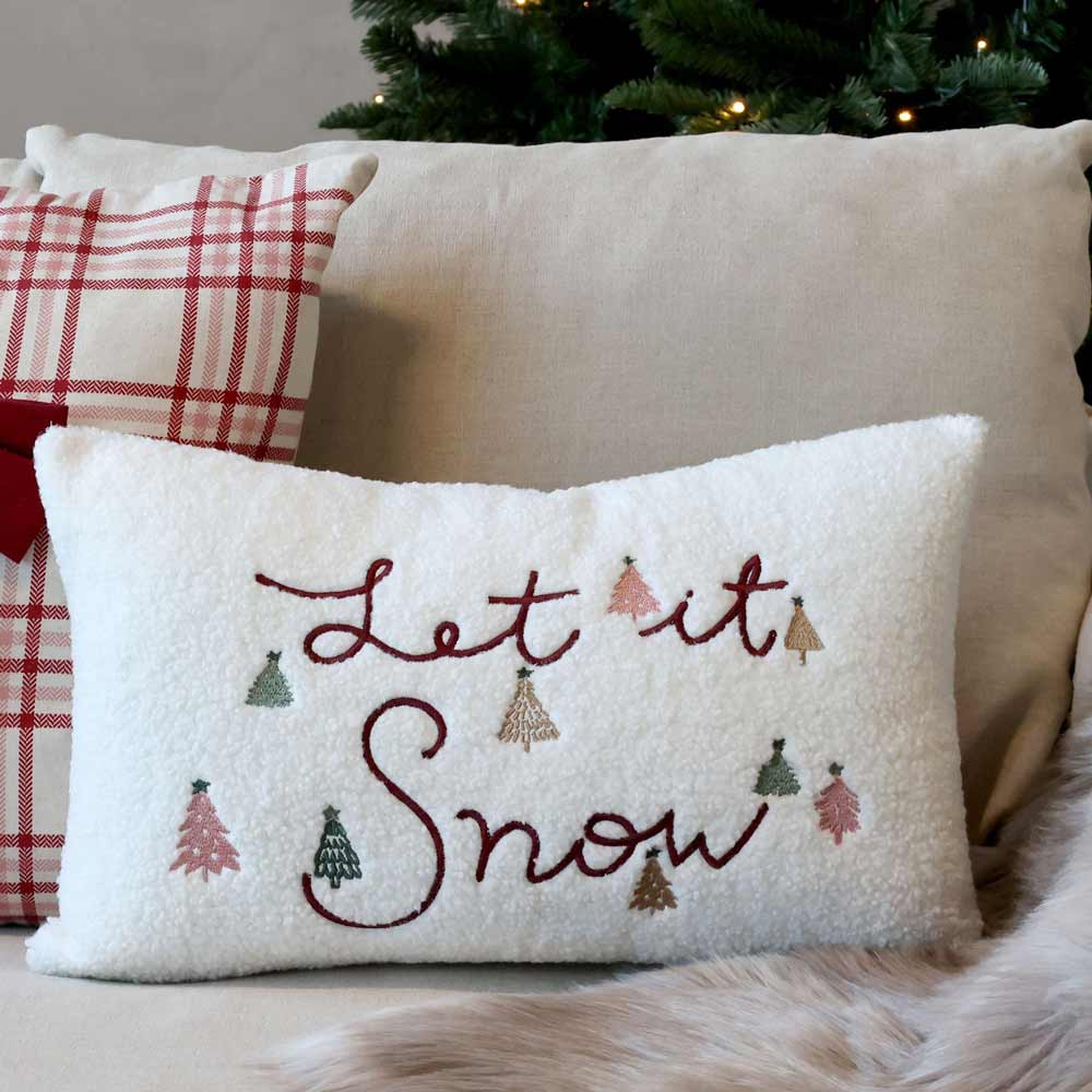 Zwei Dekokissen: eines mit rotem Karomuster und der Chic Antique Kissenbezug „Let it Snow“ in den Maßen 50 x 35 cm, verziert mit kleinen Baummotiven, liegen auf einem Sofa neben einem Weihnachtsbaum.