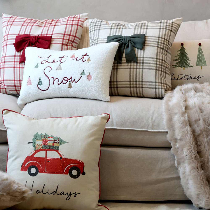 Der Chic Antique - Kissenbezug Let It Snow (50 x 35 cm) ziert ein beiges Sofa und zeigt weihnachtliche Designs mit Karomustern, Schleifen und saisonalen Motiven wie Bäumen und einem Auto mit Geschenken.