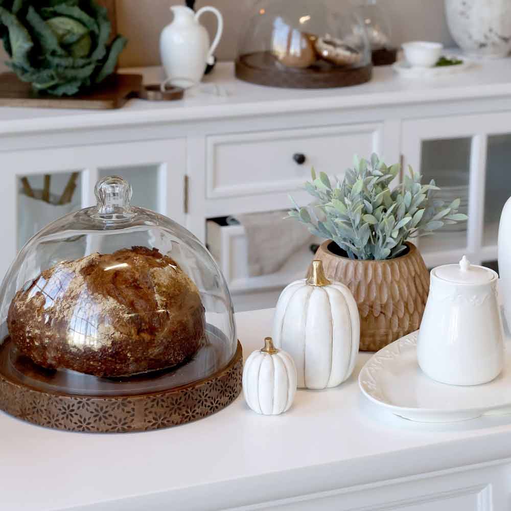Ein Laib Brot unter einer Glaskuppel steht auf einer Arbeitsplatte mit zwei Chic Antique Kürbis-Deko in Gold und Antikweiß, einer Topfpflanze und weißem Keramikgeschirr.