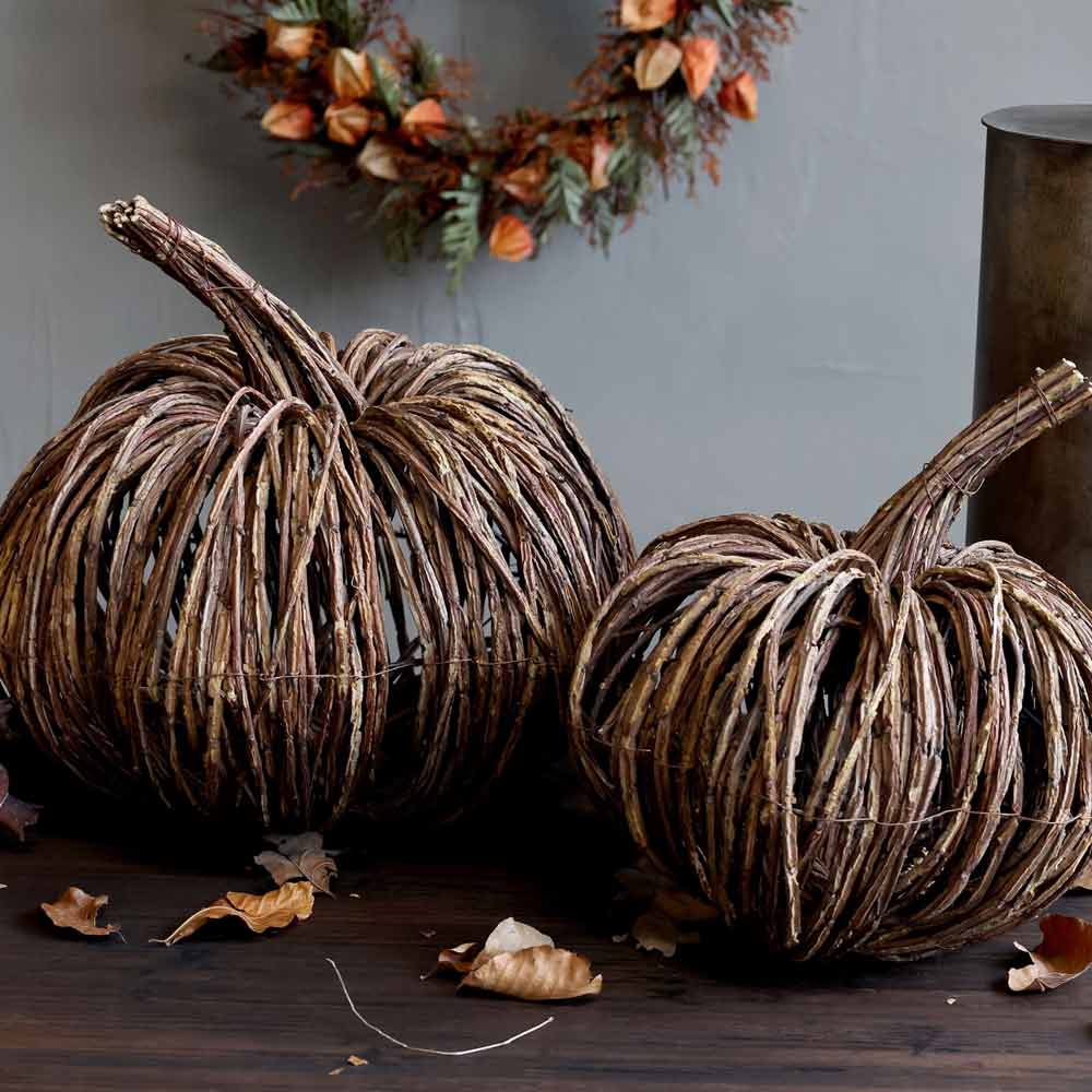 Zwei Chic Antique - Kürbis Rattan-Kürbisse aus geflochtenem Rattan liegen auf einer Holzfläche inmitten getrockneter Blätter, im Hintergrund hängt ein Kranz an der Wand.