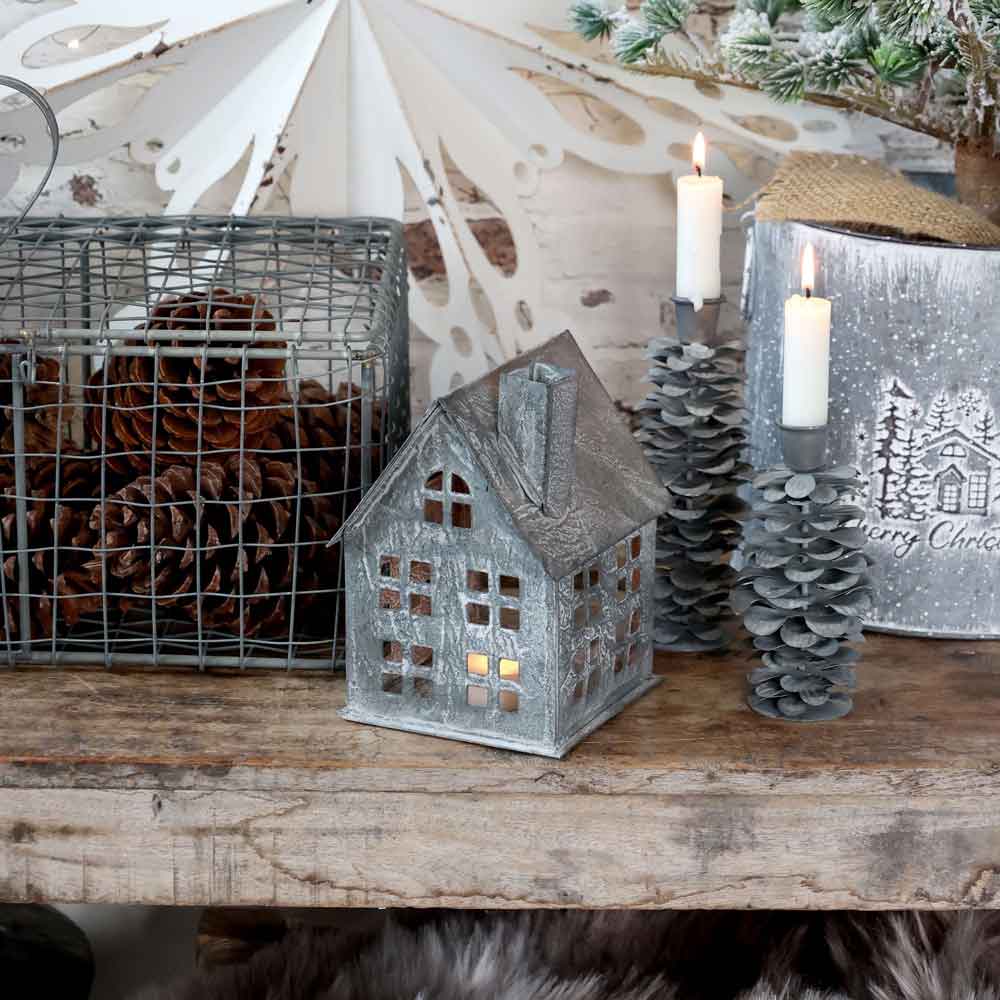 Schaffen Sie eine gemütliche Weihnachtsstimmung mit dem Chic Antique - Lichthaus Antik Zink (Höhe 17 cm), begleitet von brennenden Kerzen, einem Korb mit Tannenzapfen und einem festlichen Eimer auf einem Holztisch.