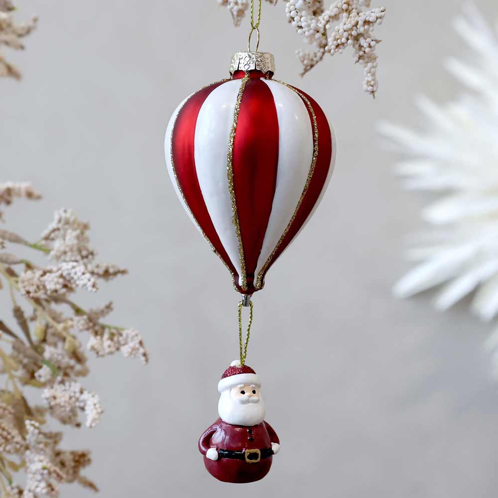 Ein Heißluftballon-Ornament von Chic Antique in rot-weißem Design mit einer kleinen, darunter hängenden Weihnachtsmannfigur, akzentuiert durch zarte dekorative Pflanzen.