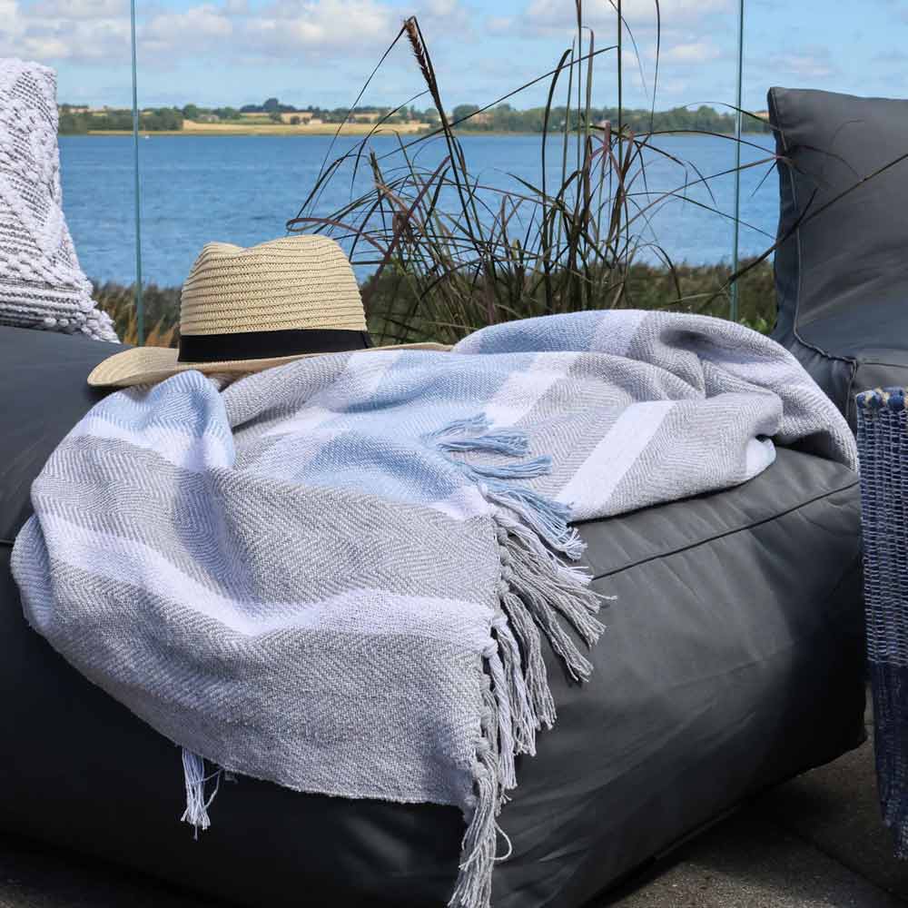Die Chic Antique - Plaid Kuscheldecke Grau-Blau mit Fransen und ein Strohhut liegen auf einer dunklen Outdoor-Liege, im Hintergrund hohes Gras und Wasser.