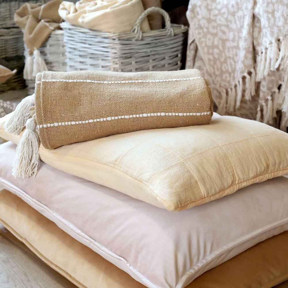 Ein Chic Antique Plaid mit Fransen im naturbraunen Boho-Look ist auf gestapelten cremefarbenen und hellgelben Kissen gerollt, im Hintergrund sind weitere Textilien und Körbe zu sehen.