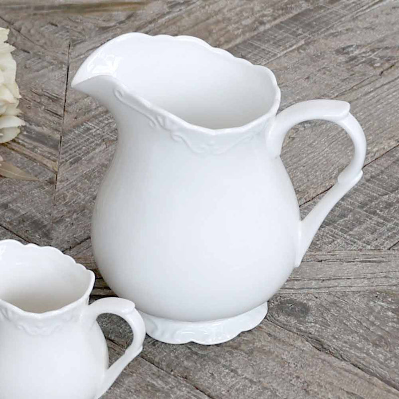 Eine große weiße Keramikkanne mit verziertem Henkel und gewelltem Rand steht auf einer Holzoberfläche neben einer passenden, kleineren Chic Antique - Provence Kanne 1 Liter.