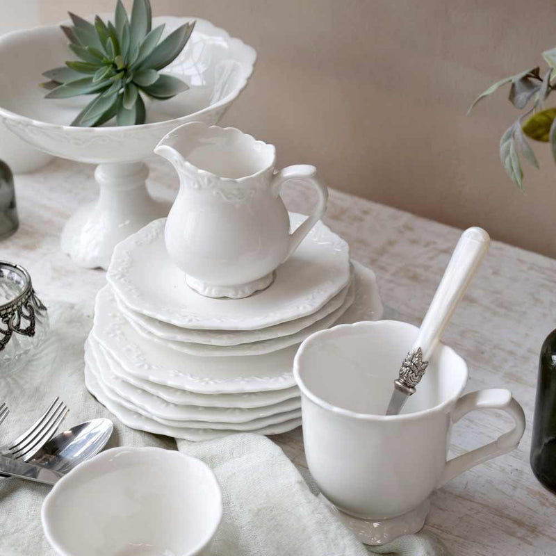 Weißes Keramikgeschirrset bestehend aus Tellern, einem Chic Antique - Provence Milchkännchen, einer Tasse mit Löffel und einer Schüssel, arrangiert auf einem Tisch mit Stoffbezug. Im Hintergrund ist auch eine Sukkulente in einer Schale zu sehen.