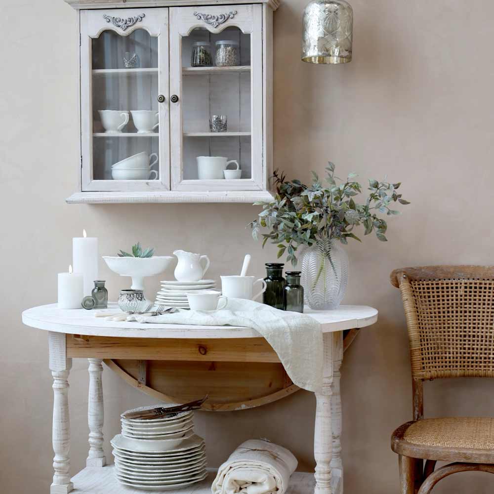 Französisch inspirierter Essplatz mit weißem Provence-Geschirr, einem Holztisch, Geflechtstuhl und verglastem Wandschrank