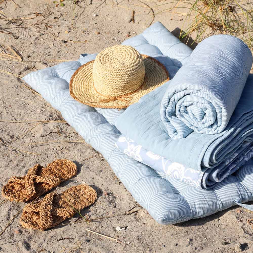 Ein Strohhut, gewebte Sandalen und der Chic Antique Quilt Maritimes Streifenmuster Sky Blue (180 x 130 cm) sind auf einem Sandstrand mit etwas Gras arrangiert.