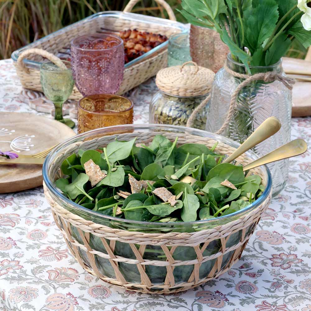 Die Chic Antique - Schüssel mit Flechtkorb ist mit Spinatsalat und Croutons gefüllt und wird auf einer gemusterten Tischdecke zwischen Tellern, Gläsern und einer Vase mit grünen Blumen platziert.