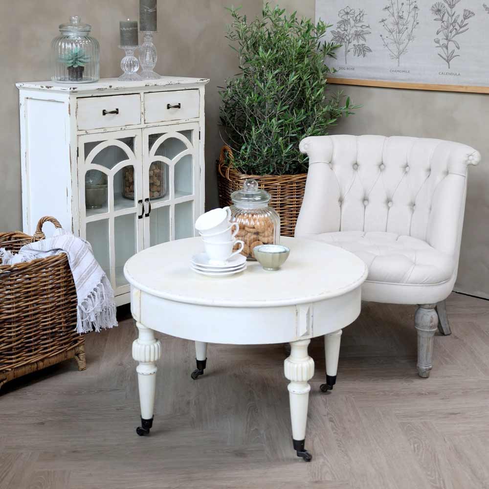 Eine gemütliche Ecke im Vintage-Stil mit einem gepolsterten weißen Sessel, einem Sofatisch Marie Antoinette mit Teeservice, einem Schrank und dekorativen Pflanzen in einem Raum mit neutralen Tönen.
