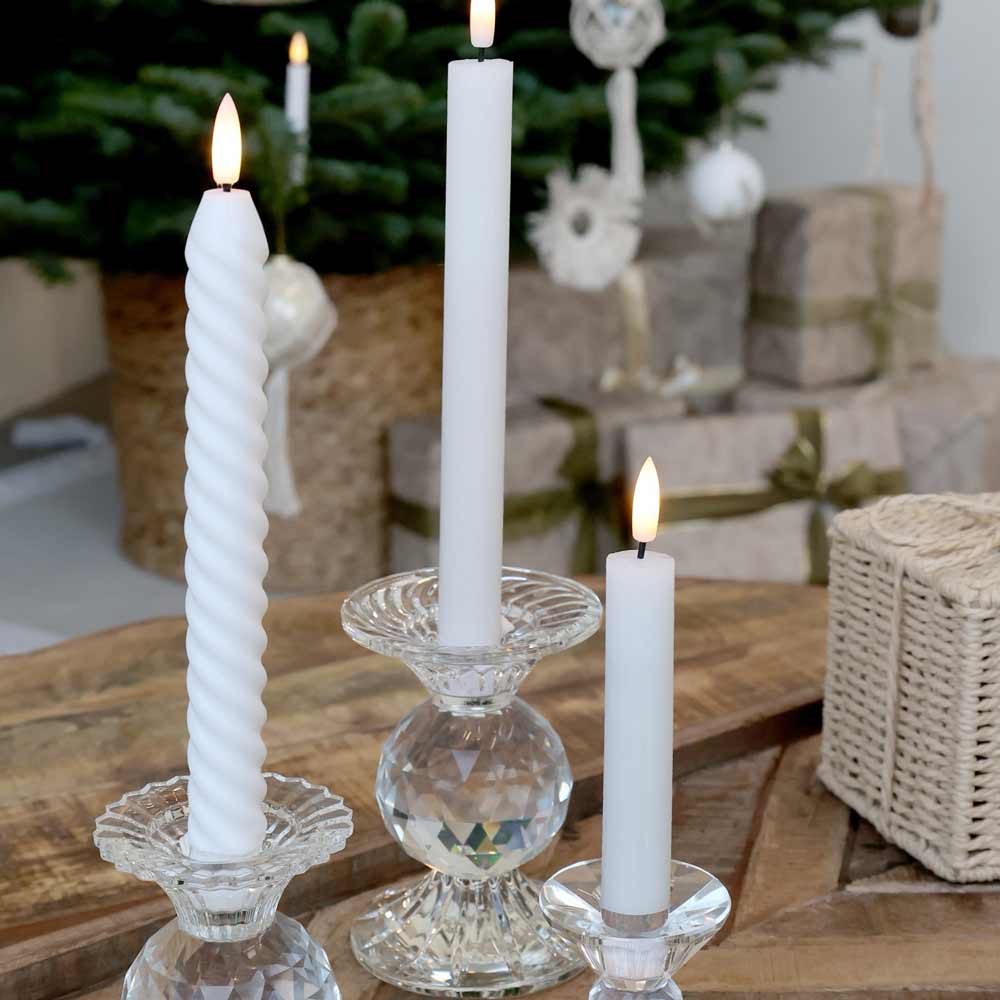 Drei Chic Antique - Stabkerzen LED 2er-Set in klaren Glaskerzenhaltern auf einem Holztisch. Im Hintergrund sind ein geschmückter Weihnachtsbaum und verpackte Geschenke zu sehen.