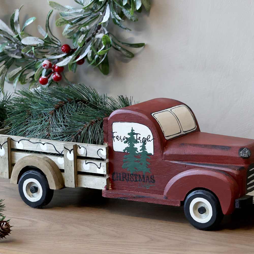 Ein Chic Antique - Vintage Auto mit Weihnachtsbaum, mit der Aufschrift „Farm Fresh Christmas Trees“ bemalt und mit immergrünen Zweigen versehen, steht auf einer Holzfläche, umgeben von weihnachtlichem Grün.