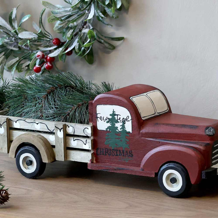 Ein Chic Antique - Vintage Auto mit Weihnachtsbaum, mit der Aufschrift „Farm Fresh Christmas Trees“ bemalt und mit immergrünen Zweigen versehen, steht auf einer Holzfläche, umgeben von weihnachtlichem Grün.