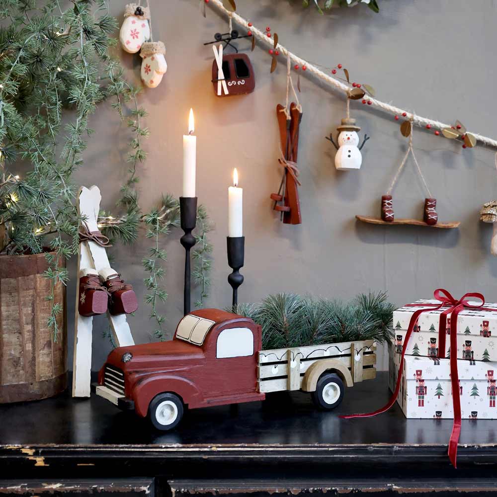 Eine festliche Szene mit „Vintage Auto mit Weihnachtsbaum“ von Chic Antique, umgeben von brennenden Kerzen und geschmückt mit Weihnachtsdekorationen, darunter Ornamente, eine Geschenkbox mit Schleife und eine an der Wand hängende Girlande.