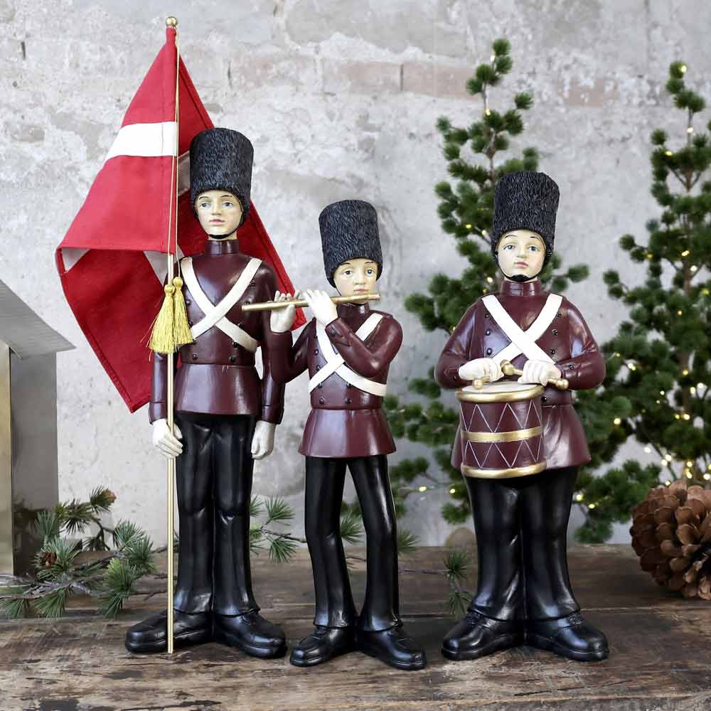 Das Chic Antique - Vintage Orchester 3er-Set besteht aus drei Spielzeugsoldatenfiguren, jeweils ausgestattet mit einer Flagge, einer Flöte und einer Trommel, die vor kleinen Weihnachtsbäumen posieren.