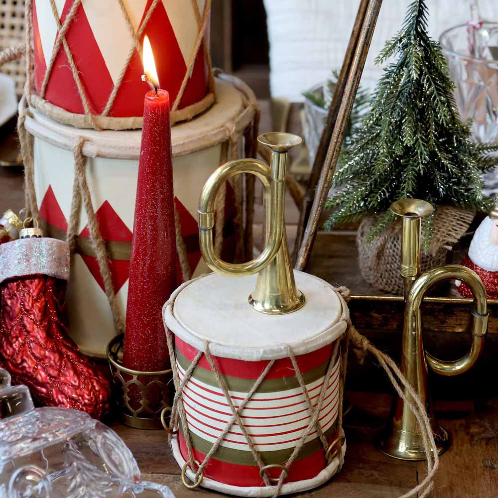 Festliche Präsentation mit einer Vintage-Trommel aus handgefertigtem Leder in Antikkirsche, neben einer roten Kerze, Messinghörnern, einem kleinen geschmückten Weihnachtsbaum und roten Strümpfen auf einem Tisch.