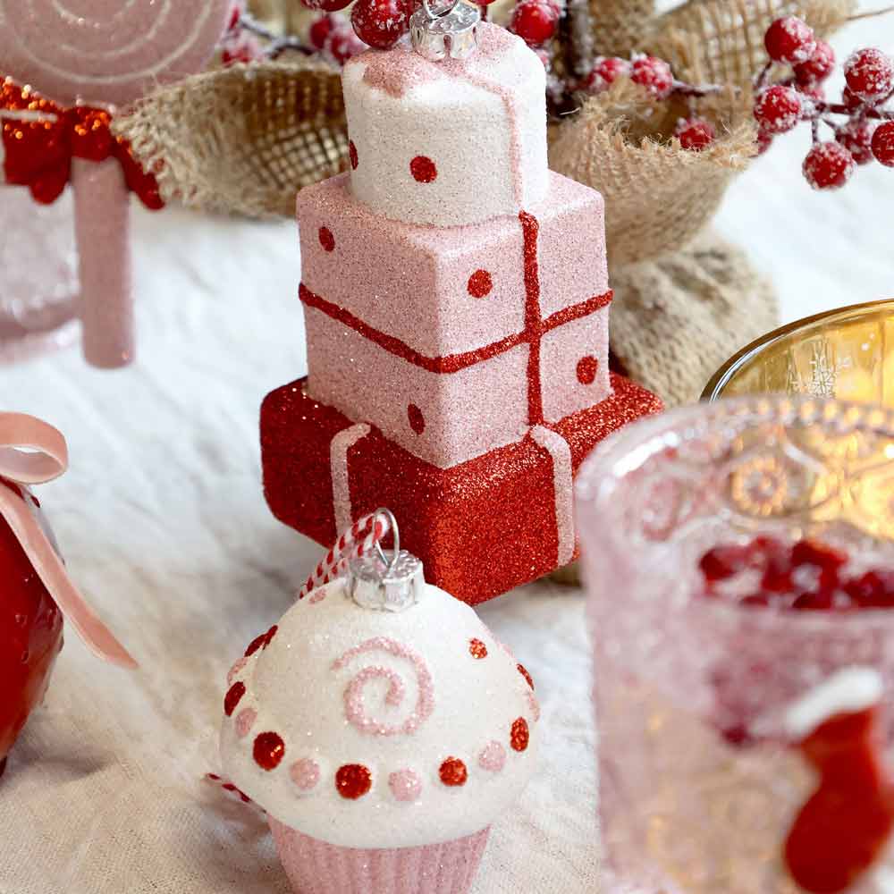 Das Chic Antique - Weihnachtskugel Candy Muffin Pink Lady-Ornament ist von festlichen Dekorationen mit gestapelten glitzernden Geschenkboxen in Rot- und Rosatönen umgeben.