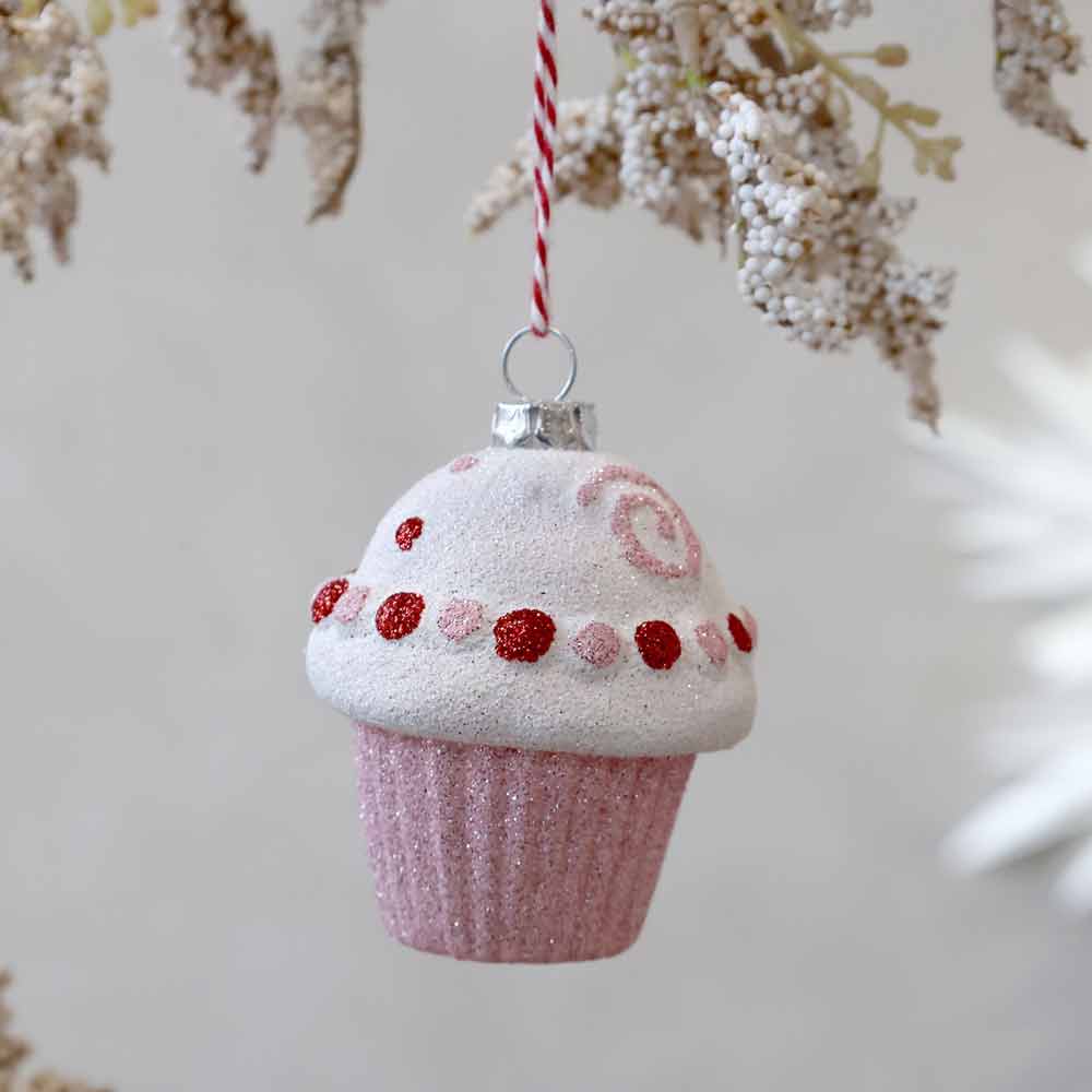 Ein Chic Antique Weihnachtskugel-Candy-Muffin-Pink-Lady-Ornament mit glitzernden Cupcake-Details mit roten und rosa Akzenten hängt an einer rot-weißen Schnur zwischen beigen Zierzweigen.