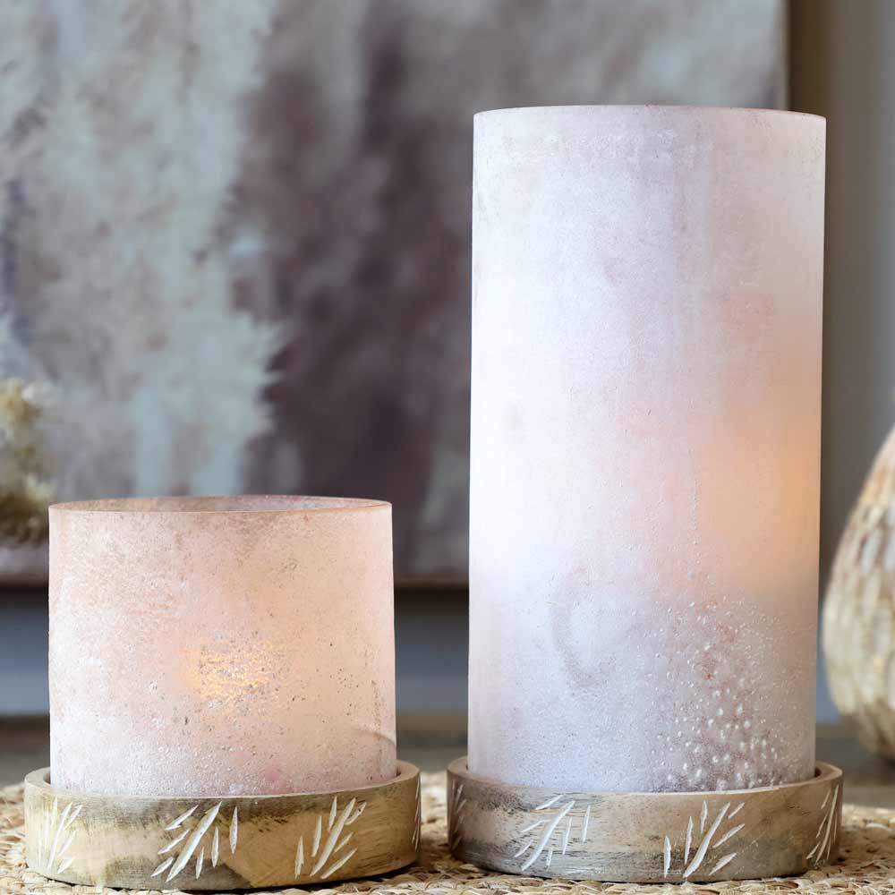 Zwei Chic Antique - Windlichter mit graviertem Sockel in unterschiedlicher Höhe stehen auf Untersetzern mit Blättermuster. Die Kerzen strahlen ein sanftes, warmes Licht aus.