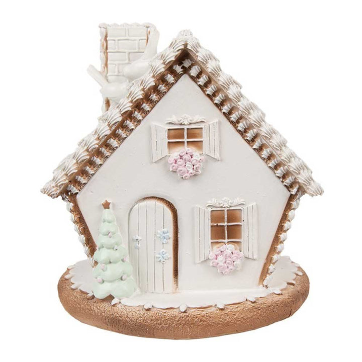 Das Clayre & Eef - Lebkuchenhaus mit LED 17 cm ist ein bezauberndes kleines Deko-Haus mit schneebedecktem Dach, von der Dachtraufe hängenden Eiszapfen, einem weißen Weihnachtsbaum davor und rosa Blumen, die die Fensterbänke schmücken.