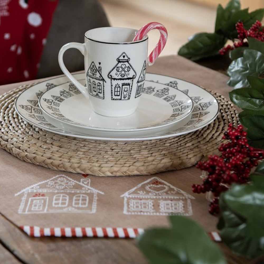 Ein weihnachtlich gedeckter Tisch mit einer Stoffserviette „Lebkuchenhäuser“ von Clayre & Eef, einer Tasse und Untertasse mit Lebkuchenhausmotiven, einem Pfefferminz-Zuckerrohr in der Tasse und festlicher Dekoration drumherum.