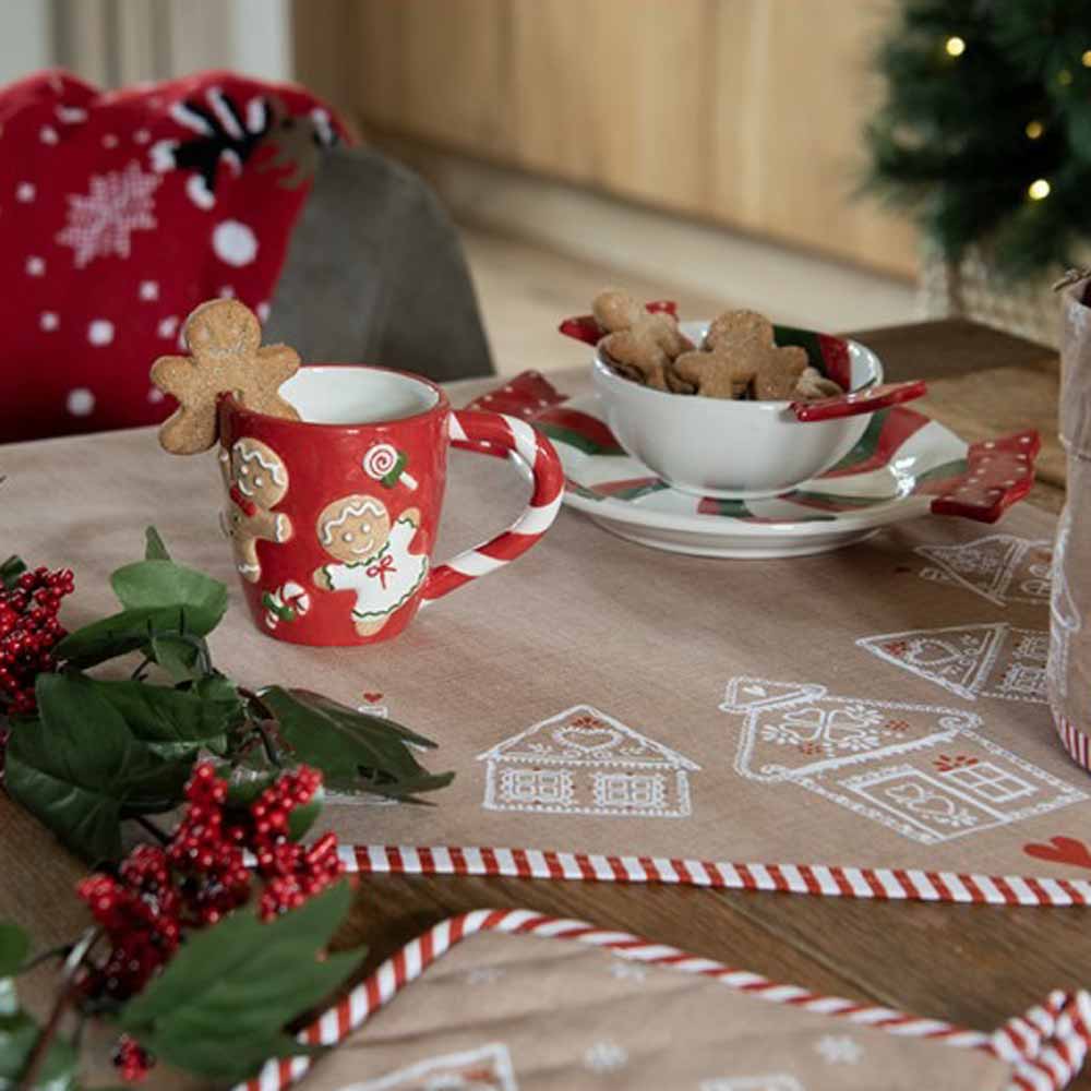 Ein festlich gedeckter Tisch mit einer roten Weihnachtstasse, einem Teller mit Lebkuchen, grünem Laub und roten Beeren. Dekoriert mit den Clayre & Eef - Stoffservietten Lebkuchenhäusern und einem kleinen Weihnachtsbaum im Hintergrund.