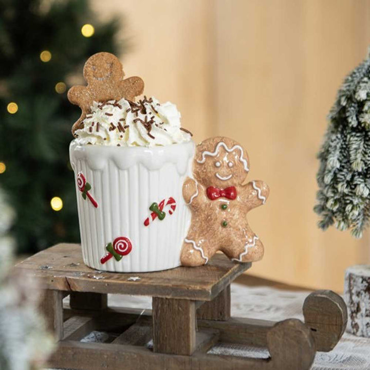 Eine festliche Pfefferkuchenmann-Tasse von Clayre & Eef, gefüllt mit heißer Schokolade mit Schlagsahne und Schokostreuseln, mit einem Lebkuchenmann-Keks im Inneren und einem weiteren daneben, alles auf einem kleinen Holzschlitten.