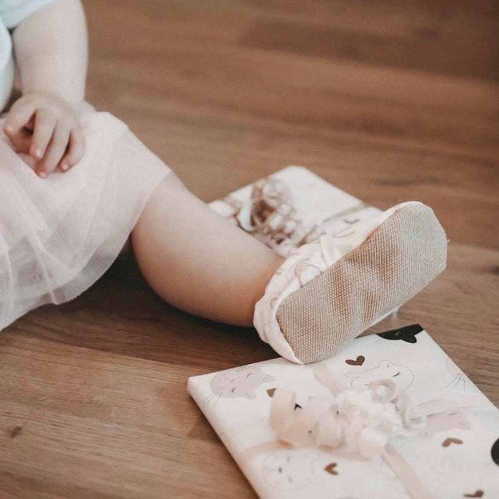 Nahaufnahme der Beine eines Kleinkindes in weißen Strumpfhosen und Ballettschuhen, das auf einem Holzboden neben einem gemusterten Kissen sitzt und Cosy Roots – Lauflernschuhe Rainbows trägt.