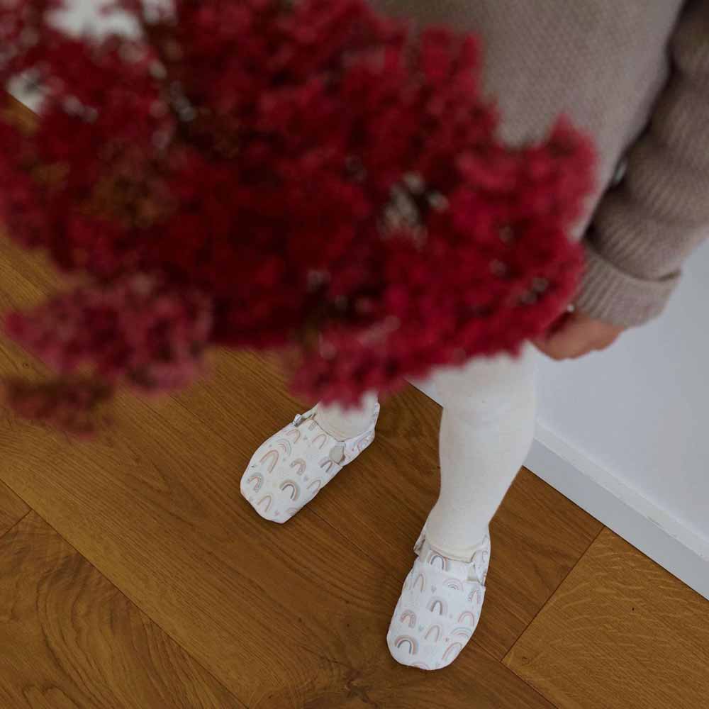 Person in Cosy Roots – Lauflernschuhe Rainbows hält einen Strauß roter Blumen und steht auf einem Holzboden.