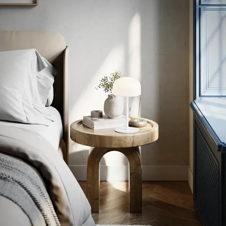 Runder Holztisch neben einem Bett, mit weißer Leuchte rechts sowie Buch und kleiner Schale auf der Tischfläche