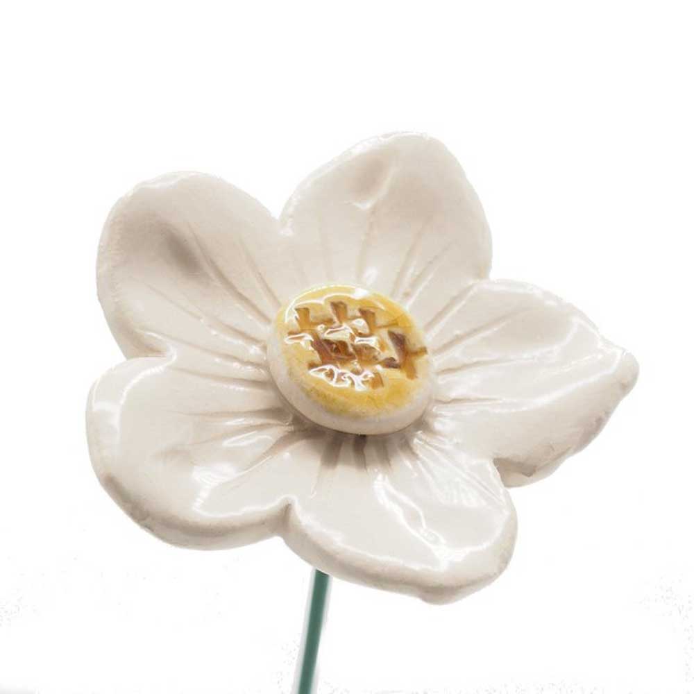 Eine Nahaufnahme einer weißen Keramikblume mit einer gelben knopfartigen Mitte und einem grünen Stiel vor einem einfarbigen Hintergrund, Abteilung M – Gartenstecker Petunie.