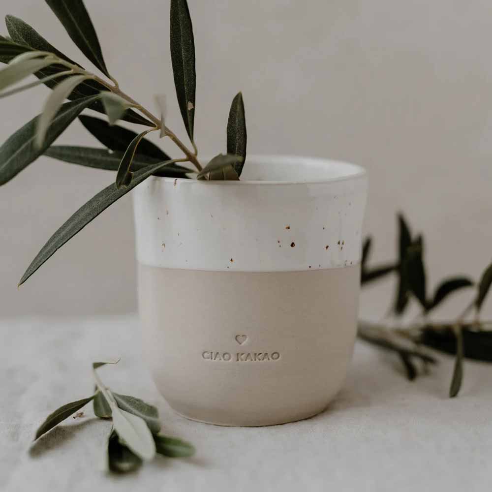 Eine Keramiktasse in neutralen Tönen mit der Aufschrift „Eulenschnitt – Becher Ciao Kakao“, in der sich ein kleiner Zweig mit grünen Blättern befindet, steht auf einer hellen Oberfläche, daneben liegen weitere Blätter.