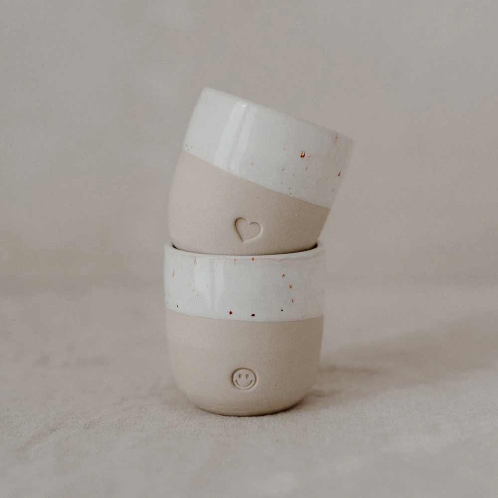 Zwei Eulenschnitt - Espresso Becher Herz Keramiktassen mit halb weißem, halb braunem Design sind gestapelt; die obere Tasse zeigt ein Herz und die untere ein Smiley-Gesicht.