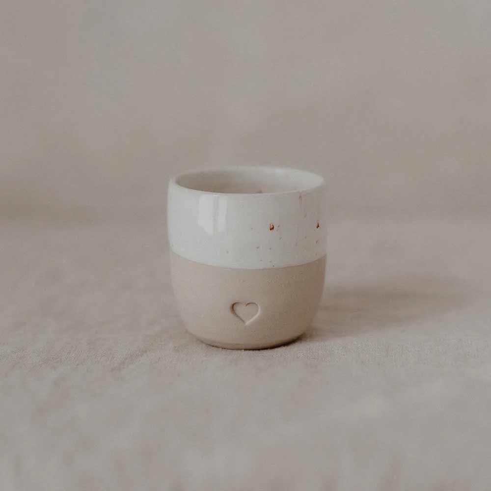 Der Eulenschnitt - Espresso Becher Herz, eine Keramiktasse mit glänzend weißem Deckel und mattbeigem Boden mit kleiner Herzvertiefung, steht auf einer neutralen Stoffoberfläche.