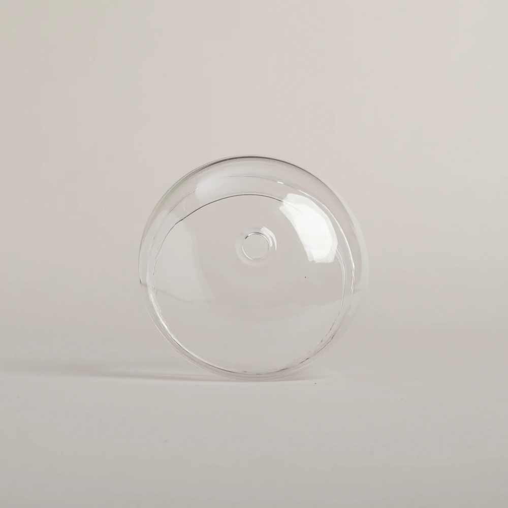 Der Eulenschnitt - Glasdeckel für Trinkglas, ein klarer, runder Glasdeckel mit einem kleinen Loch an der Oberseite, wird vor einem schlichten, hellen Hintergrund präsentiert.