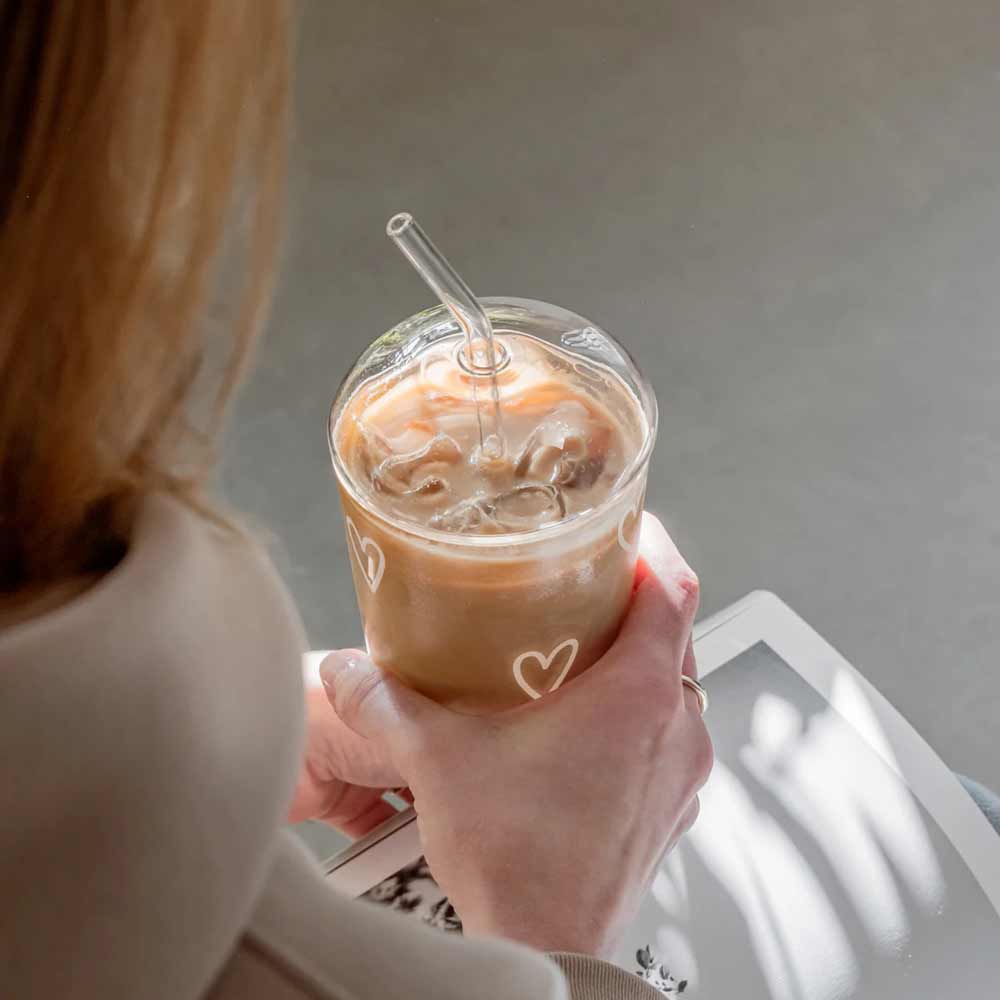 Eine Person hält einen Eiskaffee in einem Glas mit Herzmotiven, gekrönt mit dem Eulenschnitt-Glasdeckel für Trinkglas und einem durchsichtigen Strohhalm, während sie in der anderen Hand ein weißes Tablet oder Buch hält.