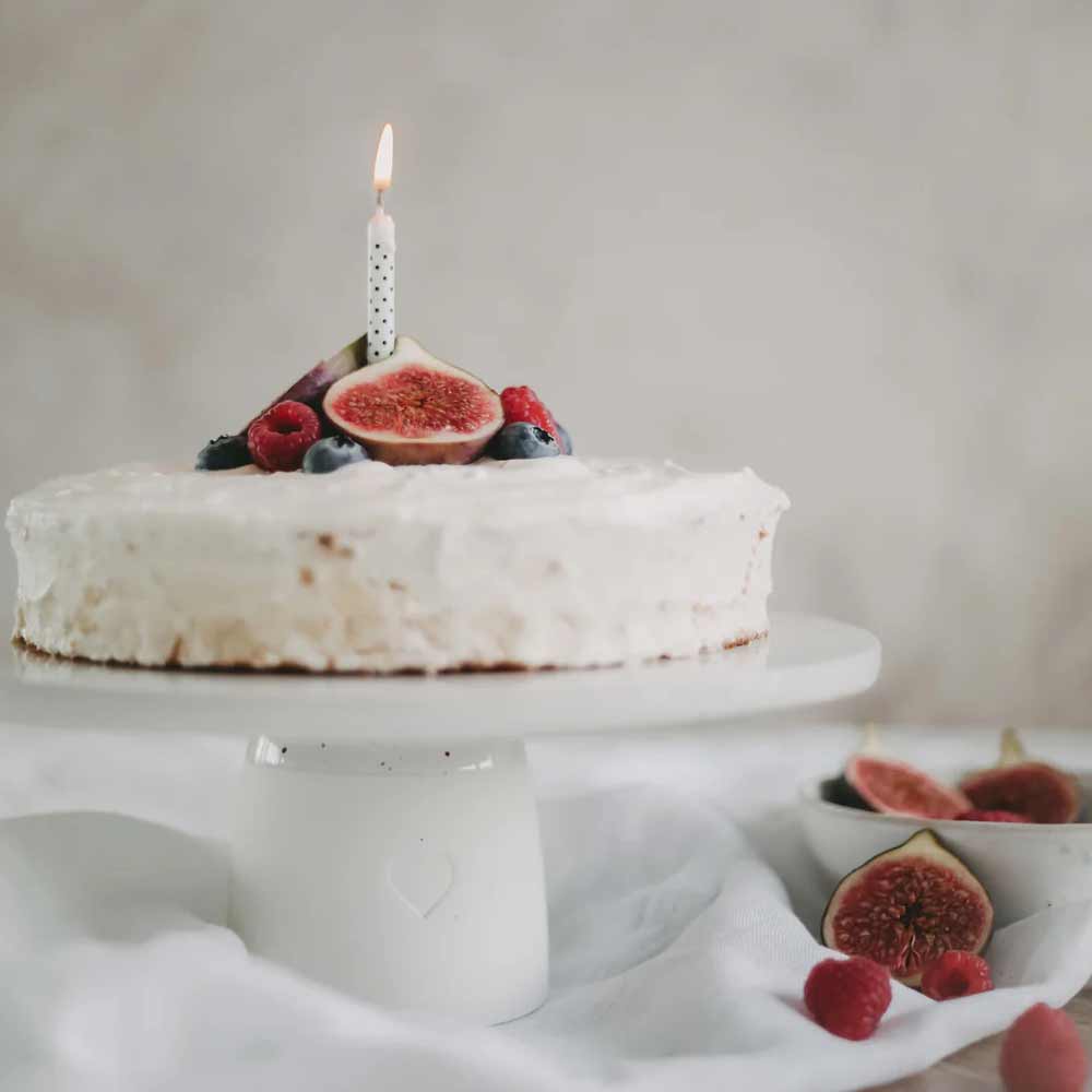 Ein runder Kuchen mit weißem Zuckerguss und frischen Feigen, Blaubeeren und Himbeeren steht auf dem Eulenschnitt - Kuchenständer Herz 10 cm mit einer brennenden Kerze. Im Hintergrund ist eine Obstschale zu sehen.