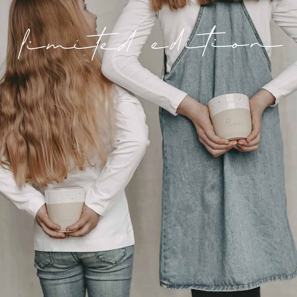 Zwei Personen mit langen Haaren und weißen Oberteilen halten Keramikbecher auf dem Rücken. Die Person rechts trägt zusätzlich eine Jeansschürze. Text: „Eulenschnitt – Limitierter Becher Mama“.