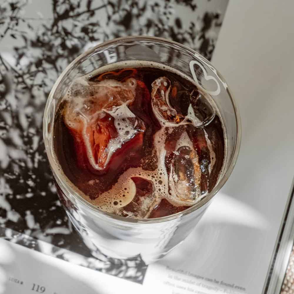 Ein Eulenschnitt - Trinkglas Herzen weiß gefüllt mit Eiskaffee und sichtbaren Eiswürfeln steht auf einem offenen Buch, das Sonnenlicht wirft sanfte Schatten.