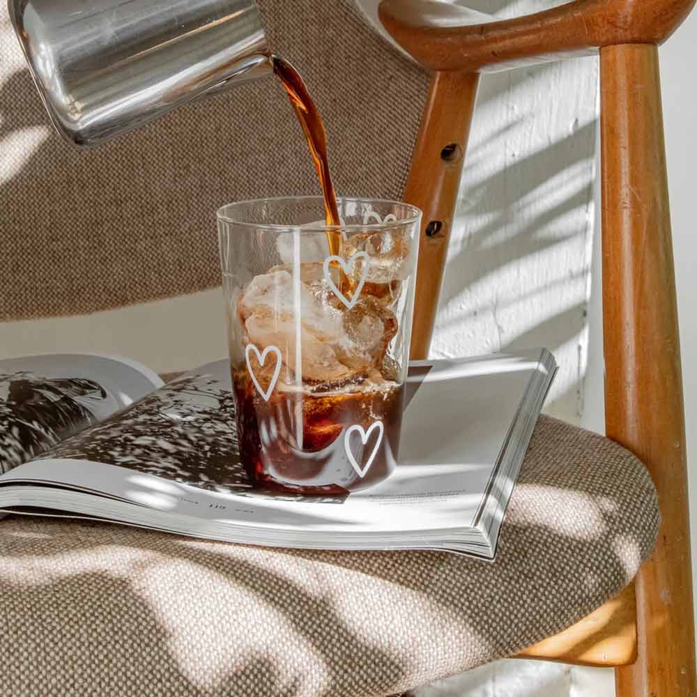 Das mit Eiskaffee gefüllte Eulenschnitt - Trinkglas Herzen weiß liegt auf einer aufgeschlagenen Zeitschrift auf einem Stoffstuhl mit Holzarmlehnen.
