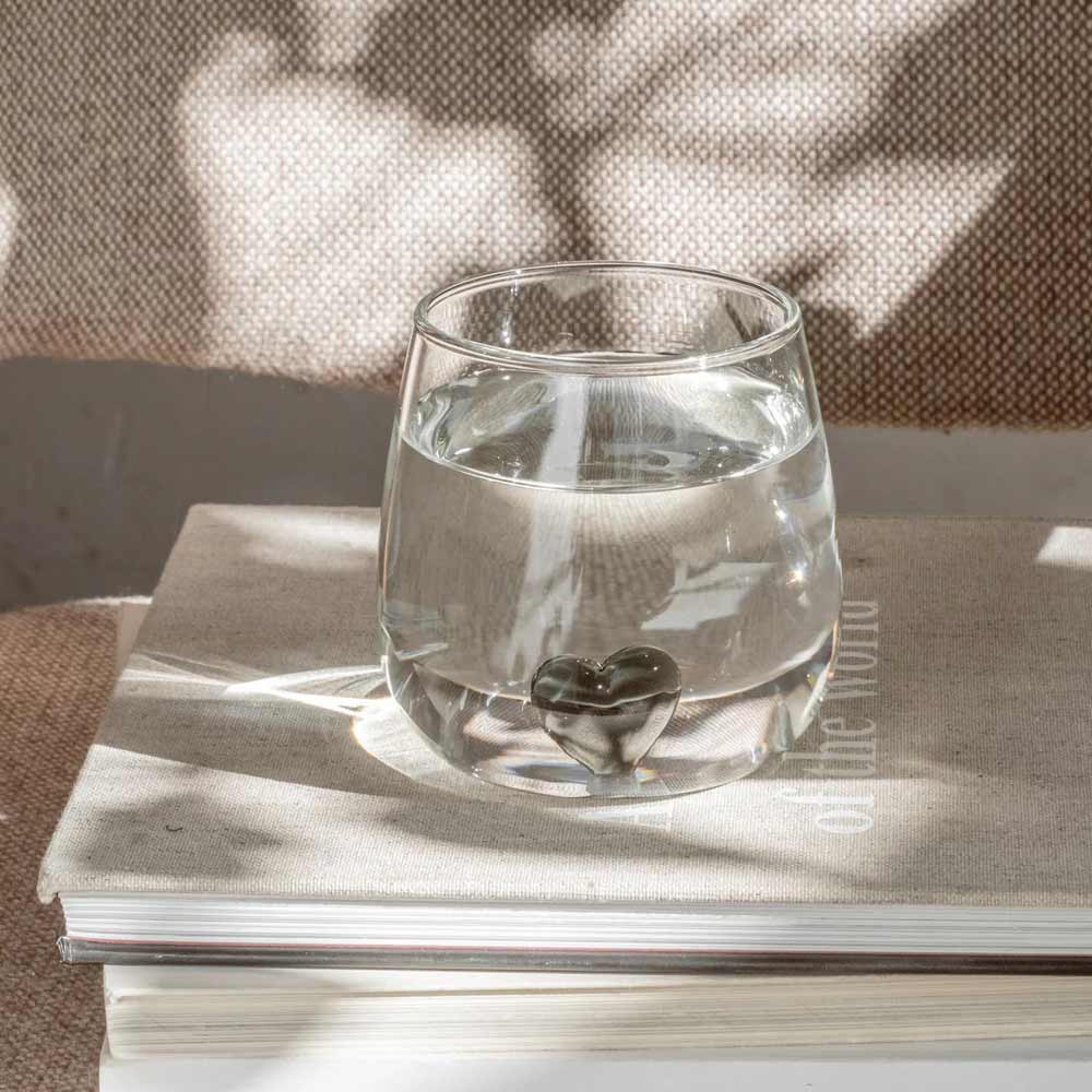 Das Eulenschnitt - Trinkglas Herzfigur schwarz mit herzförmigem Design sitzt auf einem geschlossenen beigen Buch, während im Hintergrund Sonnenlicht und Schatten spielen.