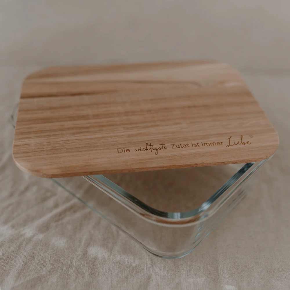 Der Eulenschnitt - Vorratsbehälter Glas mit Holzdeckel und Spruch verfügt über einen Glasbehälter mit Holzdeckel, in den auf Deutsch „Die wichtigste Zutat ist immer Liebe“ eingraviert ist, und ist auf einer beigen Stoffoberfläche abgebildet.