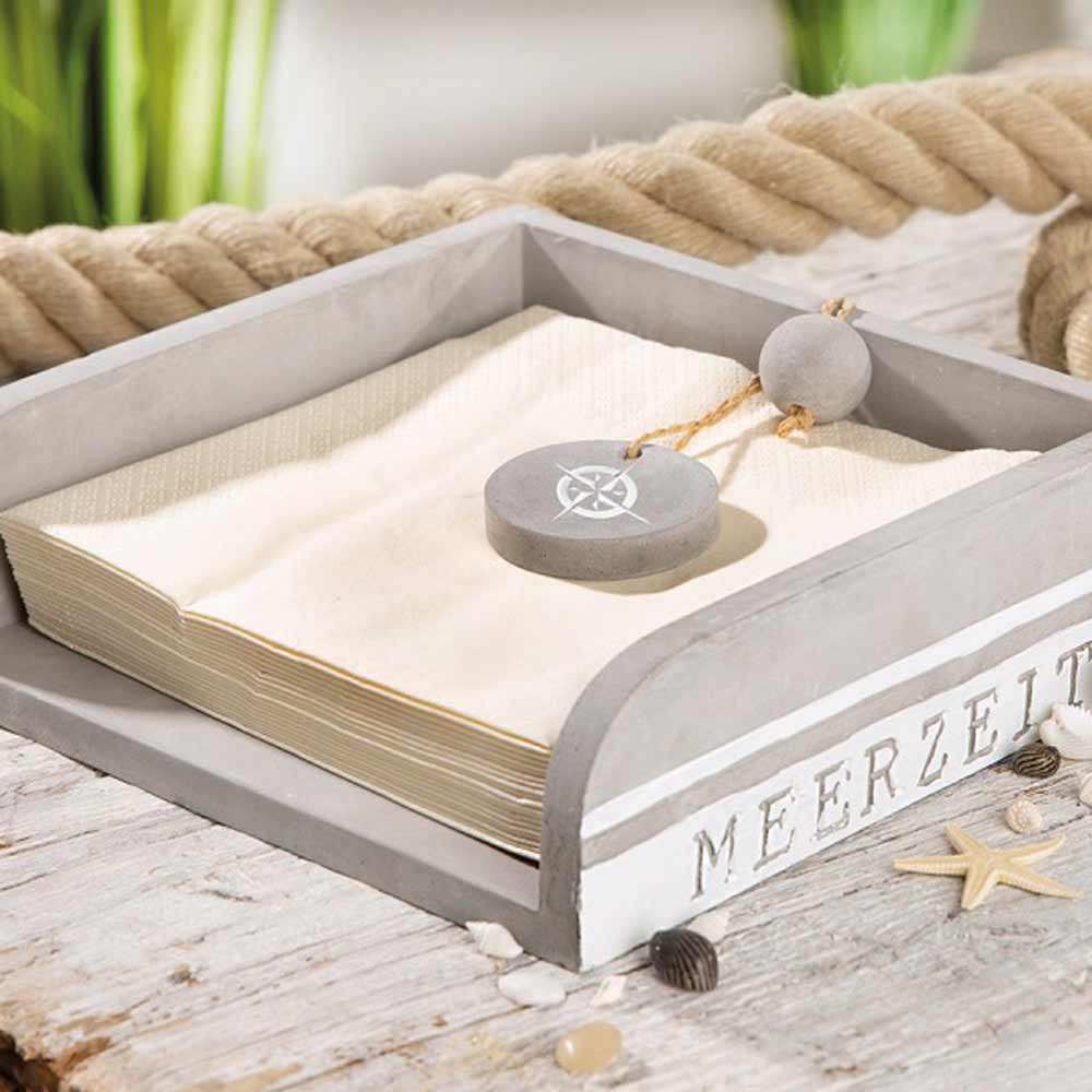 Der Gilde Serviettenhalter Meerzeit Zement in Grau hält einen Stapel beigefarbener Servietten, beschwert mit einem Stein, auf einer rustikalen Holzoberfläche, die mit Seilen und Muscheln dekoriert ist.